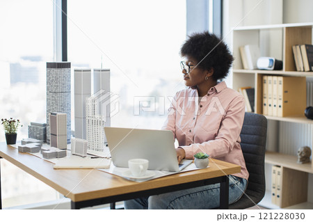 African woman architect designs modern city model. Middle-aged professional works on architectural project using laptop in bright office. Concept of creativity, innovation, urban planning. African woman architect designs modern city model. Middle-aged professional works on architectural project using laptop in bright office. Concept of creativity, innovation, urban planning. 121228030