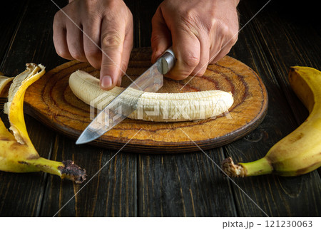 Hands skillfully slice a ripe banana on a wooden cutting board, surrounded by the remnants of other bananas. The warm kitchen atmosphere invites culinary creativity and delicious recipes 121230063