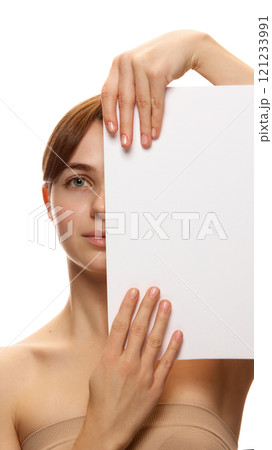 Young woman with flawless skin holding white blank paper covering half of her face against white studio background Young woman with flawless skin holding white blank paper covering half of her face against white studio background 121233991