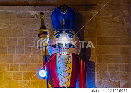 Wooden nutcracker soldier with bright red and yellow attire, illuminated by colorful blue and warm lights, stands against textured stone wall 121236971