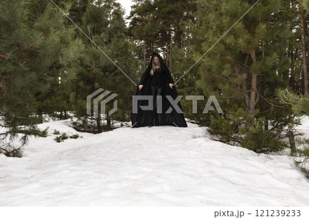 Blonde woman in a black cloak with a hood in winter in a pine forest 121239233
