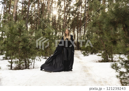 Blonde woman in a black cloak with a hood in winter in a pine forest 121239243