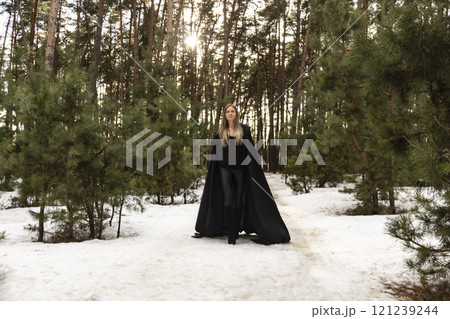 Blonde woman in a black cloak with a hood in winter in a pine forest Blonde woman in a black cloak with a hood in winter in a pine forest 121239244