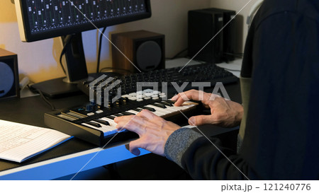 In a cozy home studio, a man focuses intently on composing music using a MIDI keyboard controller, surrounded by his musical equipment and notes, embodying creativity and inspiration 121240776