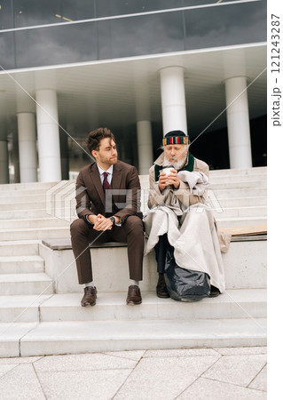 Vertical shot of kind compassionate professional in suit comforting elderly homeless man with support and kindness on building steps. Businessman showing compassion and empathy, offering support. Vertical shot of kind compassionate professional in suit comforting elderly homeless man with support and kindness on building steps. Businessman showing compassion and empathy, offering support. 121243287