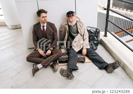 Top view of business man in suit and old homeless male sitting on piece of cardboard on ground, symbolizing social inequality and economic disparity. Concept of social issues and poverty. Top view of business man in suit and old homeless male sitting on piece of cardboard on ground, symbolizing social inequality and economic disparity. Concept of social issues and poverty. 121243289