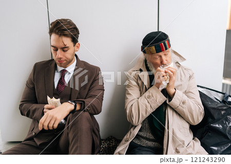 Sad businessman in suit and hungry senior homeless man sitting on piece of cardboard on city street, eating sandwiches and sharing moment of connection despite different social statuses. 121243290