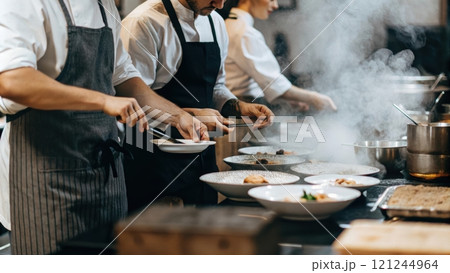 Chefs plating dishes in a busy kitchen, surrounded by steaming pots and pans 121244964