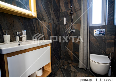 Interior of modern stylish bathroom with black tiled walls, curtain shower place and wooden furniture with wash basin and big illuminated mirror. 121245591