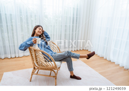 Young Asian woman happy and smiling, sitting on seat and holding mug in living room at home Young Asian woman happy and smiling, sitting on seat and holding mug in living room at home 121249039