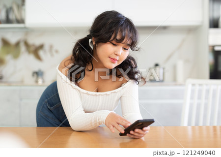 Girl standing in kitchen, leaning on countertop, and using her phone with interest 121249040