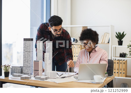 Indian man and African woman architects focused on innovative city project at desk. Collaborative teamwork in office setting with architectural models and laptop. Indian man and African woman architects focused on innovative city project at desk. Collaborative teamwork in office setting with architectural models and laptop. 121252999