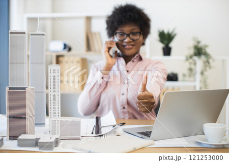Confident African businesswoman architect having phone conversation about city project. Smiling and giving thumbs up while working with laptop and architectural models in office. Confident African businesswoman architect having phone conversation about city project. Smiling and giving thumbs up while working with laptop and architectural models in office. 121253000
