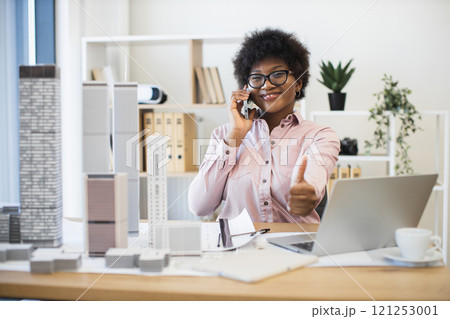 Confident African businesswoman architect having phone conversation about city project. Smiling and giving thumbs up while working with laptop and architectural models in office. 121253001