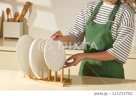 A close-up angle shows a housewife placing a plate onto a wooden holder with some plates, while a white receptacle containing some wooden kitchen utensils is placed on a beige surface. 121253702