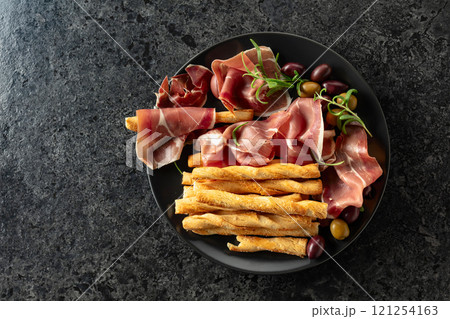 Prosciutto with bread sticks, olives, and rosemary on a black plate. 121254163