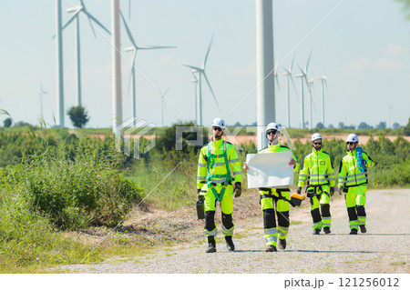 Team of professional electrical engineers in fully safety suit are working and discussing together at the windmill electric generating turbine. Electricians working at the site of windmill turbine. Team of professional electrical engineers in fully safety suit are working and discussing together at the windmill electric generating turbine. Electricians working at the site of windmill turbine. 121256012