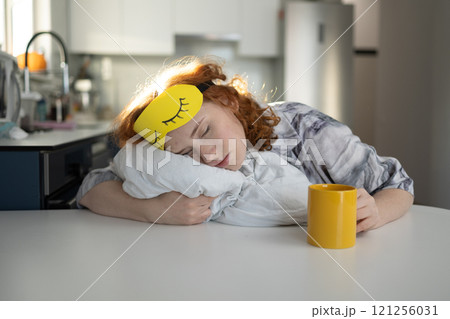 A young woman rests with a sleep mask on a table, enjoying her leisure time with a mug A young woman rests with a sleep mask on a table, enjoying her leisure time with a mug 121256031