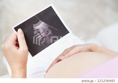 Excited and happy Asian woman looks at the ultrasound photograph of her baby inside stomach while sitting on sofa in living room at home. Asian woman is looking at ultrasound image with love. 121256485