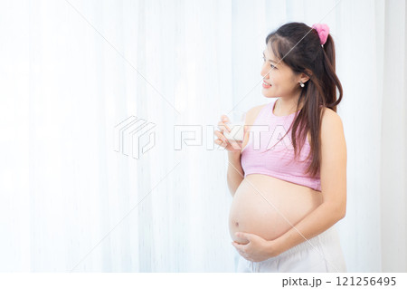 Asian pregnant woman relaxing at home.Asian pregnant woman is standing beside the window in living room and drinking a fresh milk in a glass. Asian pregnant woman portrait with copy space. 121256495