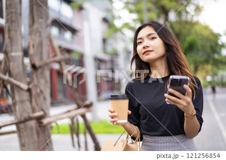 Smart and good looking Asian businesswoman is walking in the city while using a smartphone for text message and talking to colleague and other hand holding a coffee cup. Business woman portrait. Smart and good looking Asian businesswoman is walking in the city while using a smartphone for text message and talking to colleague and other hand holding a coffee cup. Business woman portrait. 121256854