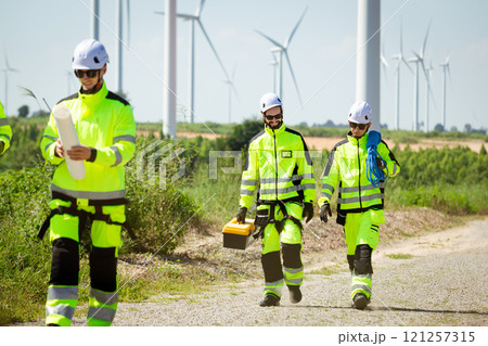 Team of professional electrical engineers in fully safety suit are working and discussing together at the windmill electric generating turbine. Electricians working at the site of windmill turbine. 121257315