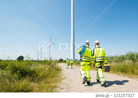 Team of professional electrical engineers in fully safety suit are working and discussing together at the windmill electric generating turbine. Electricians working at the site of windmill turbine. Team of professional electrical engineers in fully safety suit are working and discussing together at the windmill electric generating turbine. Electricians working at the site of windmill turbine. 121257327