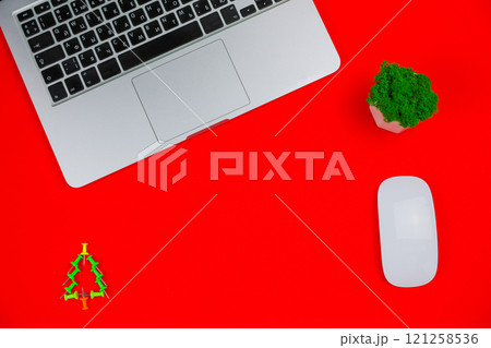 A gray laptop with a black keyboard lies on top of a photo on a red background, next to it are a creative Christmas tree consisting of green pushpins, a white computer mouse, a green plant in a white 121258536