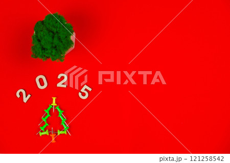 A creative Christmas tree consisting of green stationery pushpin on a bright red background, above it are wooden numbers 2025 and a green plant in a white pot with space for text. The concept of a 121258542