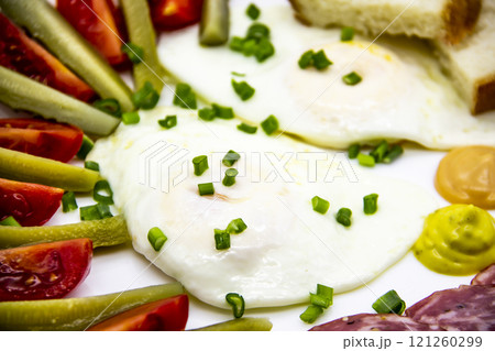 Delicious breakfast with eggs, salami, cucumbers, cherry tomatoes, slices of loaf, sauces and green onion on top on a white plate. Healthy and tasty meal for breakfast or lunch. 121260299