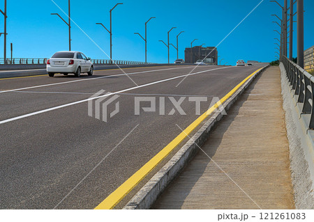 Six-lane highway on which cars move, with white dividing lines and yellow indicating edge of roadway against blue sky. Sidewalk is fenced with metal railings. empty space, free road, road markings Six-lane highway on which cars move, with white dividing lines and yellow indicating edge of roadway against blue sky. Sidewalk is fenced with metal railings. empty space, free road, road markings 121261083
