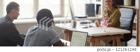 Website header shot of young African American man using laptop with white black screen in English class talking to teacher, while sitting at desk in language course group Website header shot of young African American man using laptop with white black screen in English class talking to teacher, while sitting at desk in language course group 121261248