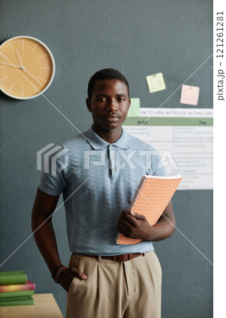 Vertical portrait shot of happy mixed raced male student posing with exercise book in hands while standing against blue wall in school classroom, copy space 121261281