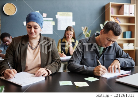 Medium shot of adult Muslim woman writing in exercise book sitting at desk with male fellow student writing test while studying English in language course group, copy space 121261313