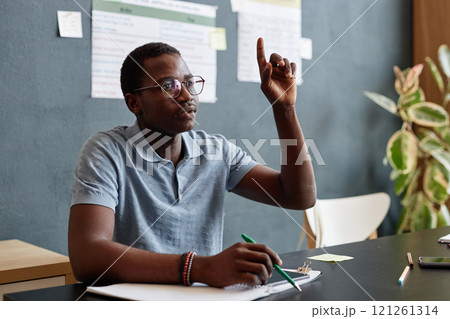 Medium shot of African American young man raising hand in class asking question while sitting at desk attending English course i n language school, copy space Medium shot of African American young man raising hand in class asking question while sitting at desk attending English course i n language school, copy space 121261314