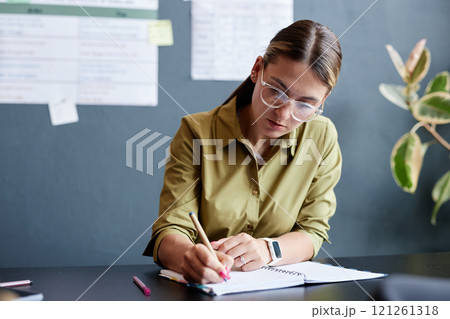 Medium shot of young concentrated female teacher writing in notebook grading students papers while sitting at desk in classroom of English language school, copy space 121261318