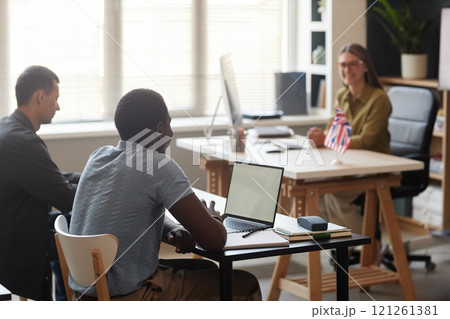 Rear view of young African American man using laptop in English class talking to female teacher while sitting at desk in classroom, copy space 121261381
