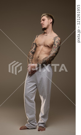 Full-length image of handsome young man with tattooed muscular body, posing shirtless in gray pants against beige studio background 121261955
