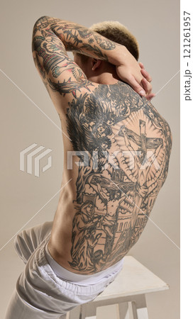 Back view of male athletic male body with tattoos, posing shirtless on chair against neutral studio background 121261957