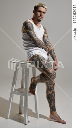 Full-length portrait of handsome young man with blonde hair, tattooed body, posing in underwear and tank top against grey studio background 121262013