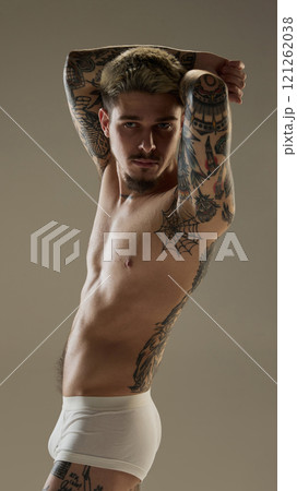 Portrait of handsome young guy with hands raised, posing shirtless in underwear against minimalistic studio background. Tattooed body 121262038