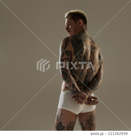 Rear view of tattooed male model with hands crossed behind his back, showing athletic tattooed body and strong physique in studio setting. 121262056