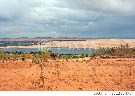 The dramatic Mui Ne sand dunes with sparse vegetation and a lake under a cloudy sky in Vietnam The dramatic Mui Ne sand dunes with sparse vegetation and a lake under a cloudy sky in Vietnam 121262479