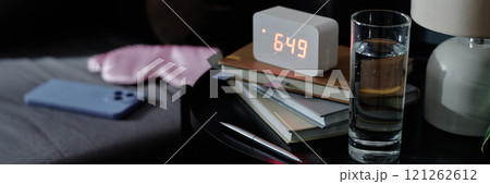 Website banner of nightstand with glass of water, books and alarm clock on top next to bed with sleep mask and smartphone 121262612