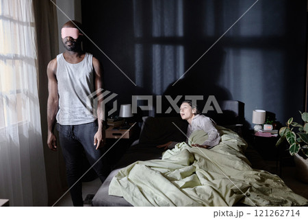 African American male sleepwalker standing next to window while his wife lying in bed and trying to help him 121262714