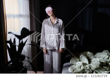 Sleepy woman dressed in crinkly cotton pajamas and pink sleep mask standing between bad and window illumed with moon light through window 121262739