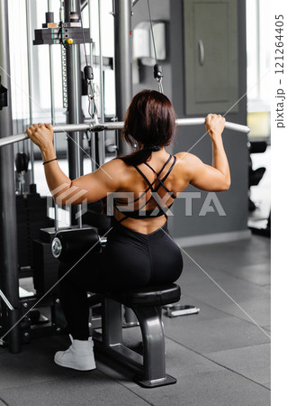 Muscular athletic female bodybuilder in black suit pulled on sports simulator in gym. Back muscles are very tense. Muscular athletic female bodybuilder in black suit pulled on sports simulator in gym. Back muscles are very tense. 121264405