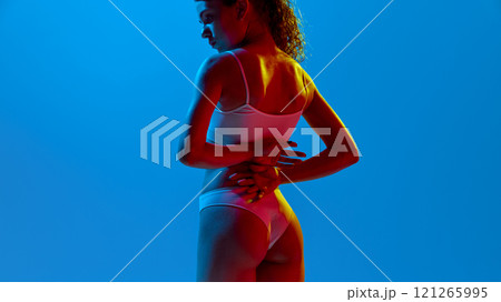 Rear view photo of young attractive woman posing in lingerie holding hand behind back in neon light against gradient background. 121265995