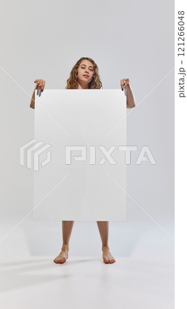 Happy, young attractive woman with curly hair posing holding poster with negative space for your ad against grey studio background. Happy, young attractive woman with curly hair posing holding poster with negative space for your ad against grey studio background. 121266048