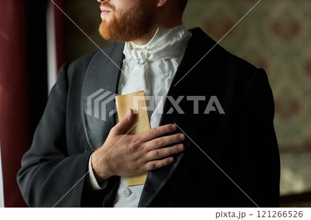 Close up of red haired man in formal attire holding letter by chest while standing in soft light by window, copy space 121266526
